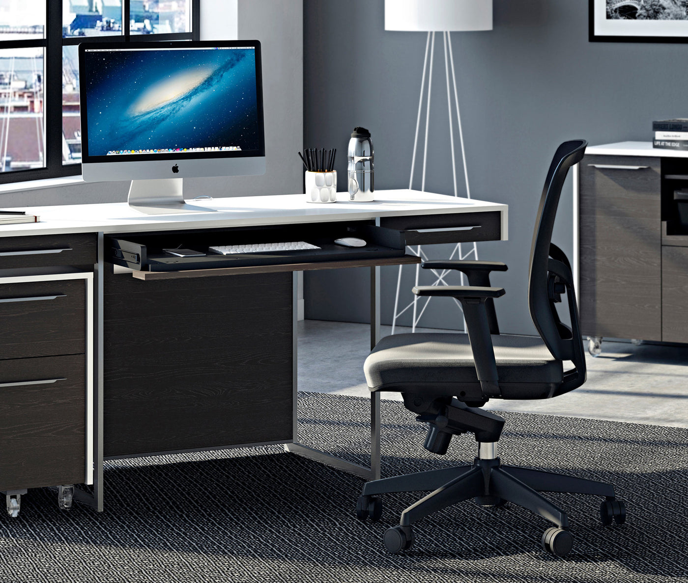 Lifestyle image of an office with a modern looking desk and mac on top and a rolling office chair slightly pulled out from it