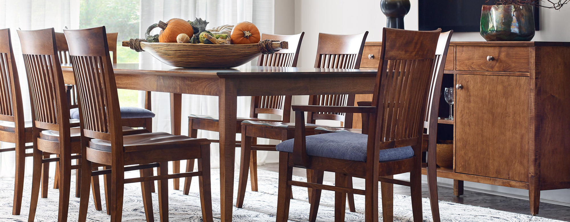 Lifestyle of a Gable Road Dining Table with a large basket full of gourds on top of it. Eight matching chairs surround the table and there is a Gable Road Large Server behind it against a white wall.