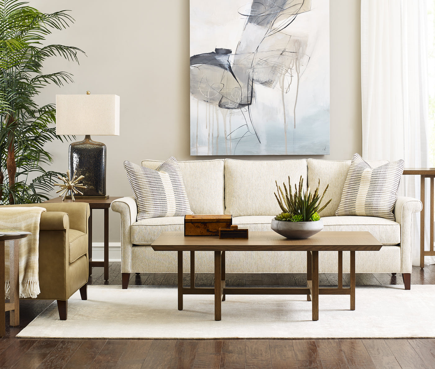 Origins by Stickley Belleville living room upholstery set up, showcasing a white fabric sofa and tan leather chair with a coffee table in front of them. There is a large white, gray, and blue painting behind the sofa and a green plant next to the sofa.