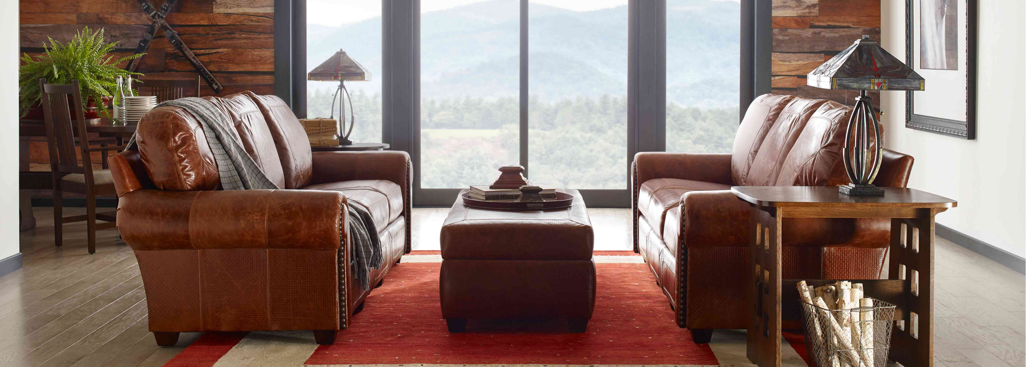 Two brown leather Santa Fe Sofas sit across from each other with a matching Santa Fe Ottoman between them, there is a large glass window in the background showing mountains outside