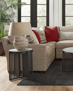 Lifestyle of a light brown fabric 7000 Series Sectional that has brown and red accent pillows. There is a Park Slope round end table next to the sectional with a found base lamp on the end table