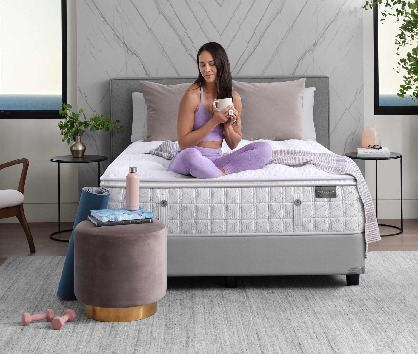 A woman wearing a light purple yoga outfit sits on top of a bare Aireloom mattress, there is a gray wall behind the bed and two windows on either side