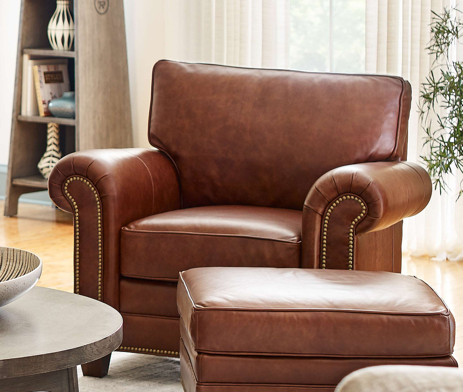 Lifestyle of a reddish brown leather Malden Chair sitting in front of a large glass window, the image shows off the nailhead that is the defining feature of the Malden line.