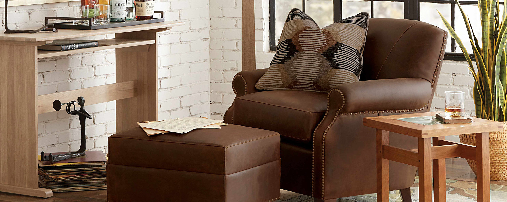 Lifestyle of a dark brown leather Beacon Club Chair with a matching Beacon Ottoman. There is a Mission Tile-Top End Table next to it that has a drink on top of it