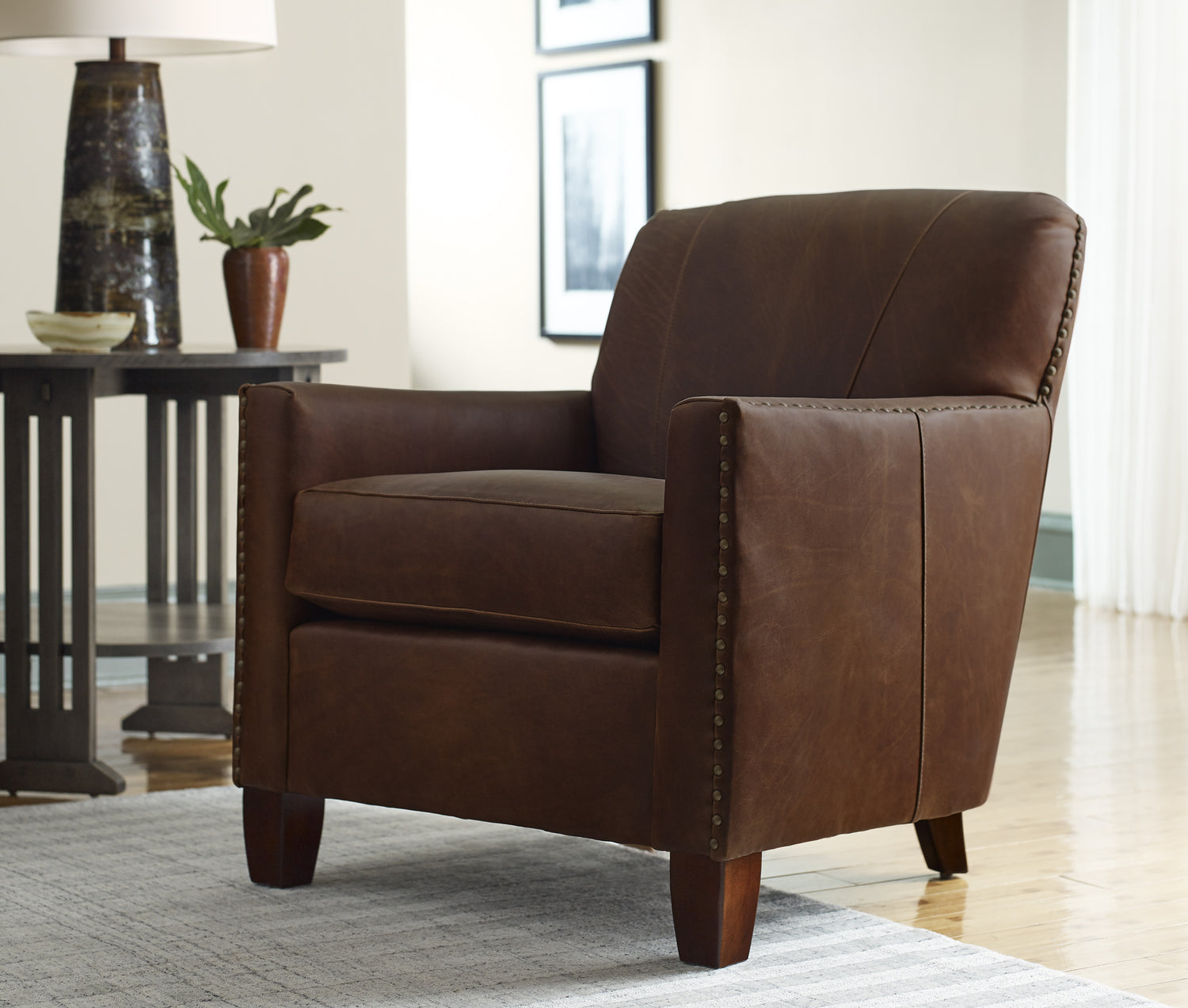 Lifestyle of a dark brown leather Santa Cruz Club Chair sitting in a white room filled with natural sunlight from the window. There is an end table next to it in the background with a tall black table lamp on top of it as well as a small brown vase.
