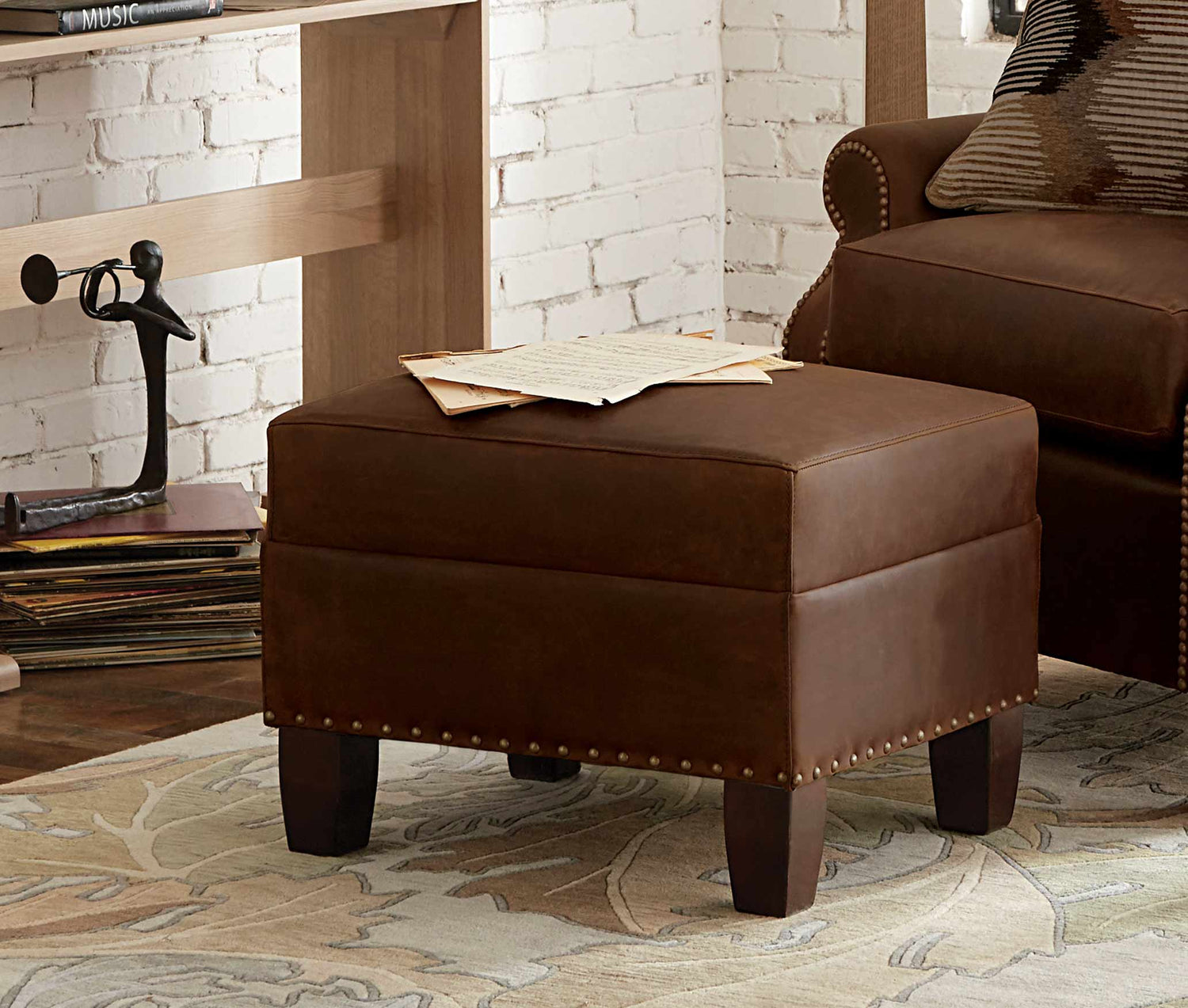 Lifestyle of a dark brown leather Beacon Ottoman that is sitting on top of a flora patterned rug. There are two small magazines sitting on top of the ottoman.