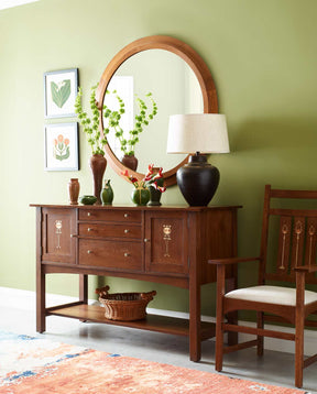 Lifestyle of a Harvey Ellis sideboard against a green wall with decorative vases on top and a round, wooden mirror above it and a matching chair to the right.