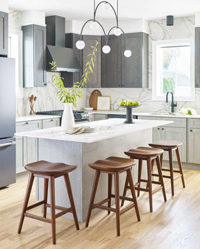 Four Walnut Grove Counter Stools surrounding a white kitchen island that has a white vase full of greenery, and a stand with green apples on top.