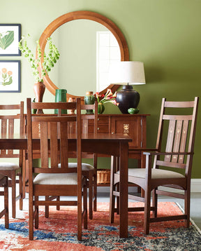 A Harvey Ellis dining room table surrounded by matching chairs with light gray upholstered seats, there is a matching sideboard in the background against a green wall with a wooden circular mirror.