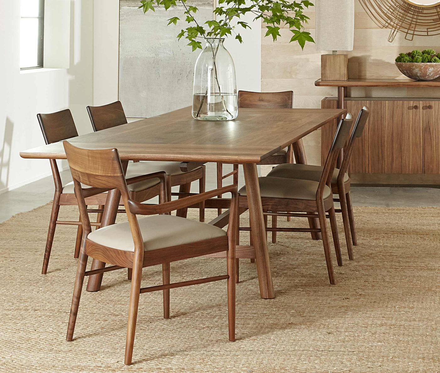 A Walnut Grove dining table surrounded by matching chairs that have white upholstered seats, there is a sideboard in the background and a clear vase full of greenery on the table.
