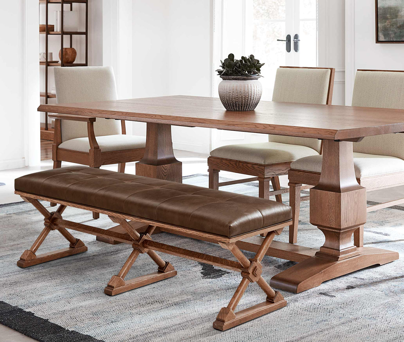 A St. Lawrence Trestle dining table with three cream upholstered chairs and a bench with a brown leather seat lining one side of the table.