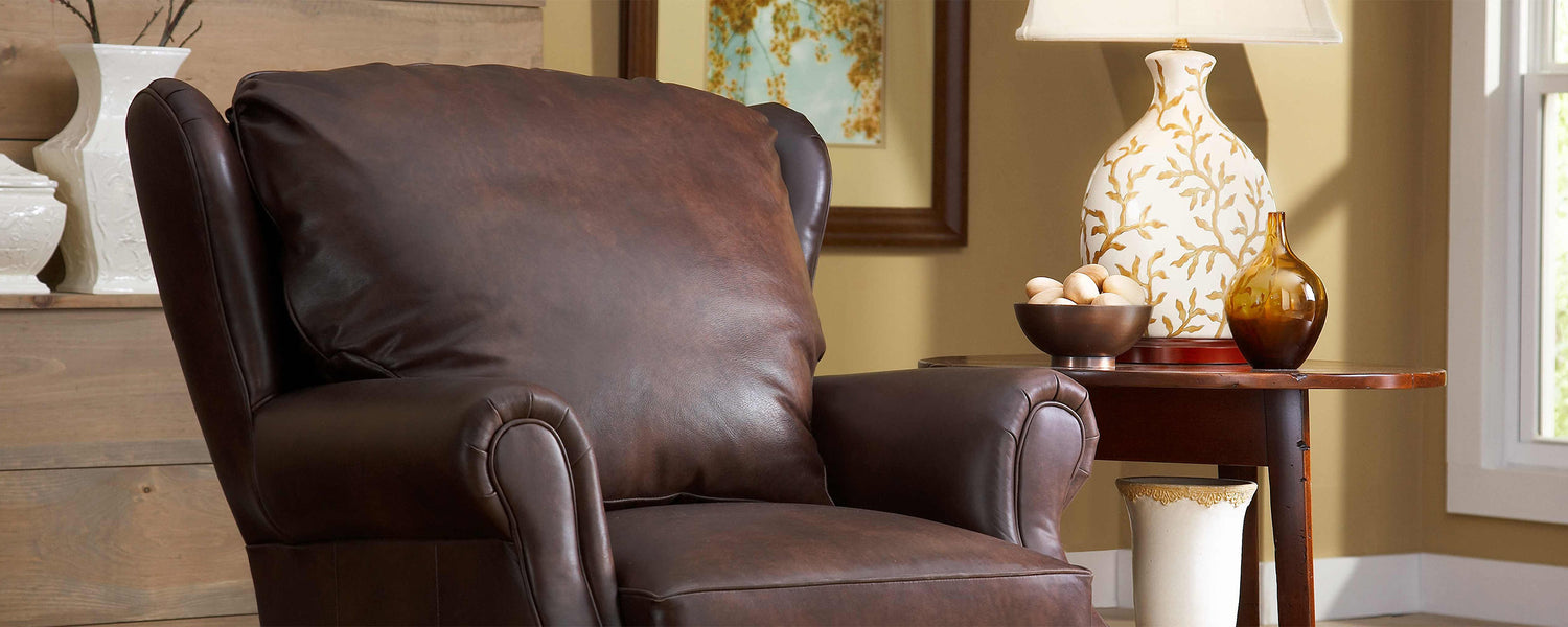 Lifestyle of a dark brown leather Grisham Recliner. It sits in a tan room with a wooden end table sitting next to it that has a floral lamp and two small decorative vases on top