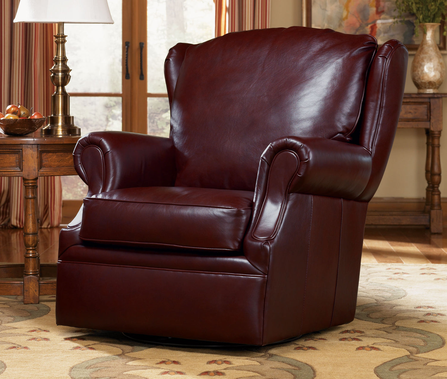 Lifestyle of a dark brown leather Grisham Swivel Chair that sits on a floral printed rug with a wooden end table beside it that has a gold lamp and basked of apples on top of it.