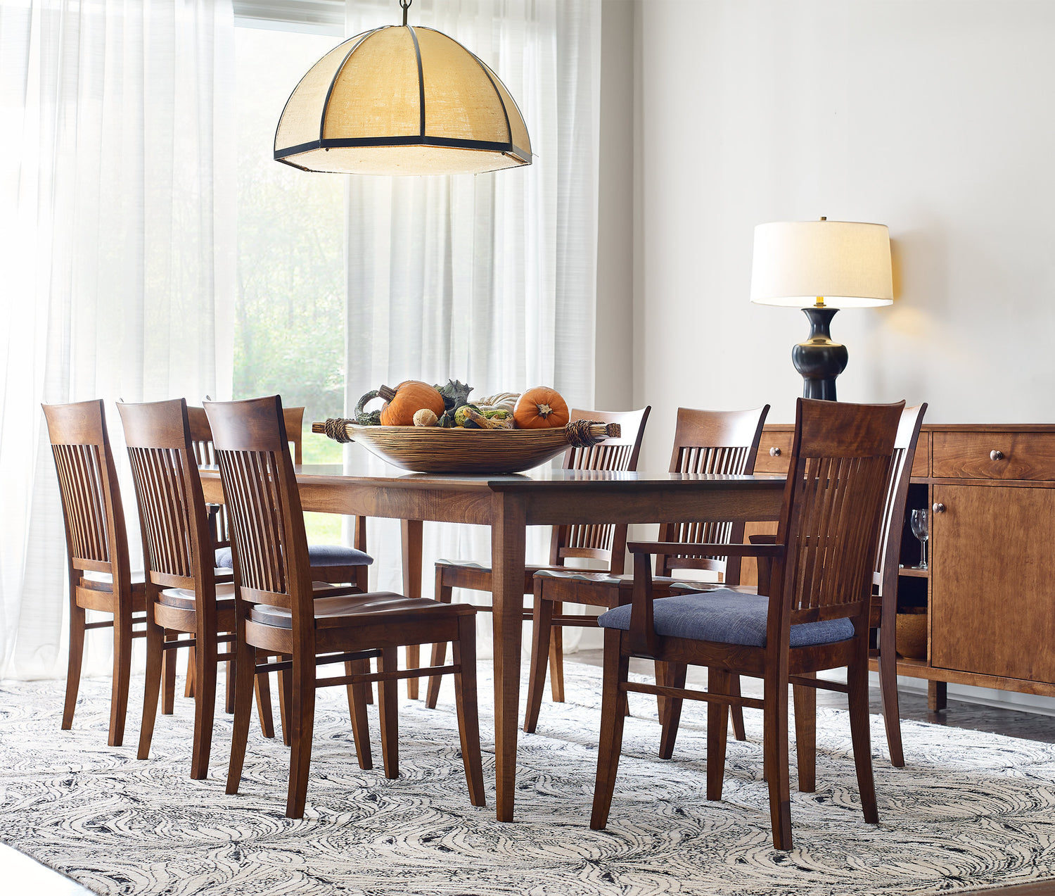 A well-lit dining room featuring a Gable Road Dining Table surrounded by eight Gable Road Chairs with dark blue upholstered seats. A decorative bowl filled with pumpkins and gourds is at the center of the table. Above the table hangs a pendant light with a yellow shade. A Gable Road Large Server with simple, straight lines and a classic table lamp with a white shade is in the background near sheer curtains.
