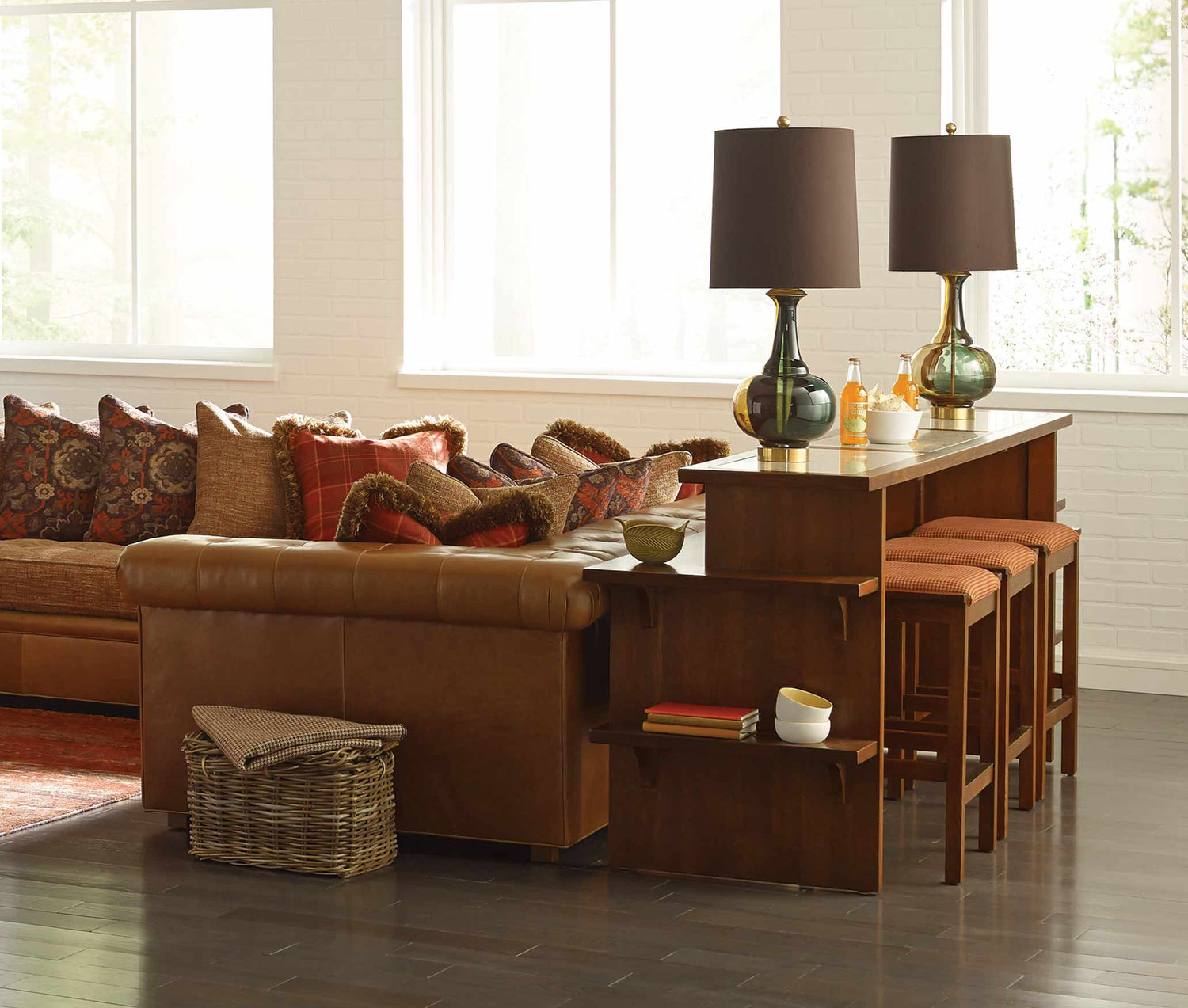 Lifestyle of a Gathering Island with matching stools against a brown leather sectional. The Gathering Island has two matching lamps on top of it as well as two drinks.