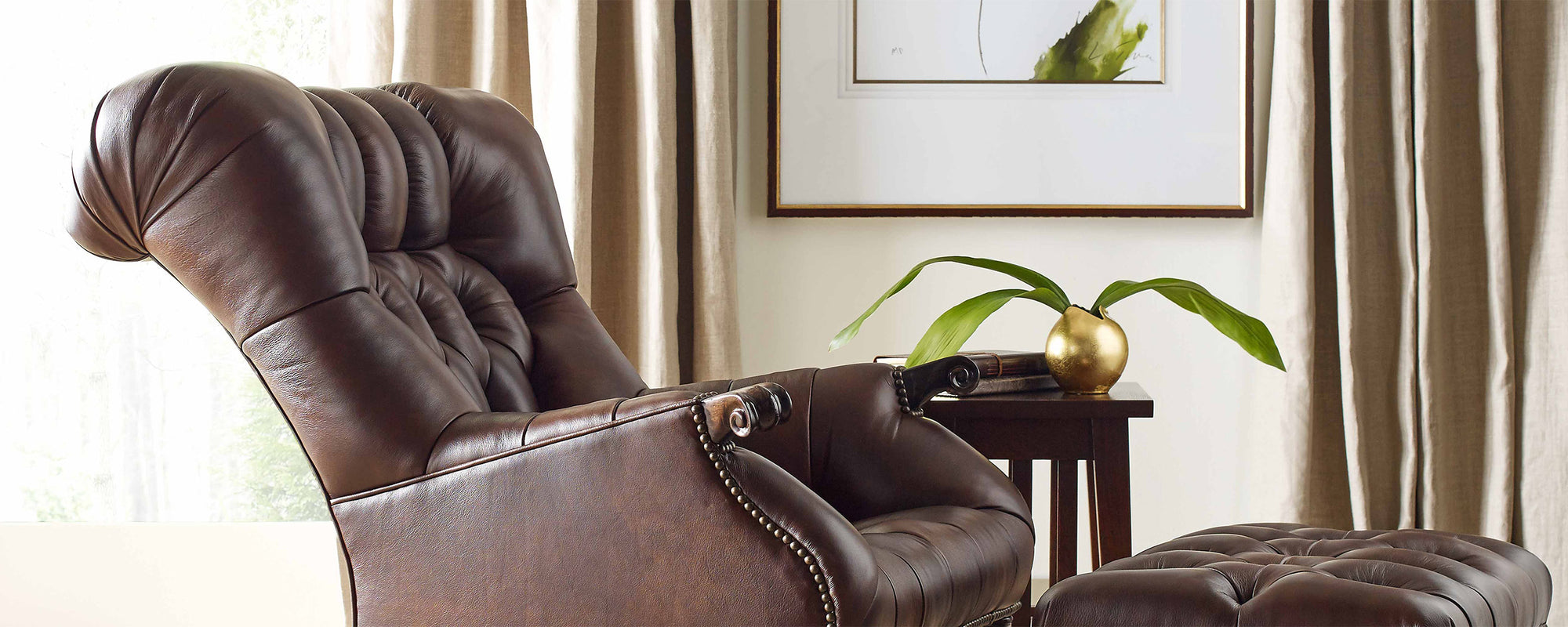 Lifestyle of a Leopold's Chair and ottoman sitting in front of a cream wall with beige curtains lining two windows. There is an end table next to the chair with a small, round gold vase