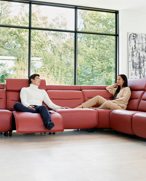 Two people relaxing on a red leather Stressless sectional in front of 3 floor to ceiling windows