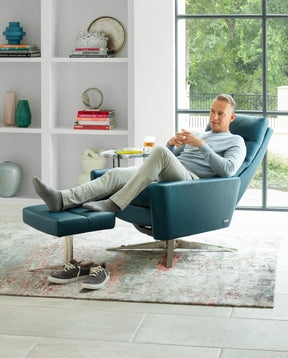 A man relaxing on a blue American Leather chair and ottoman in front of a white tall shelf and floor to ceiling window