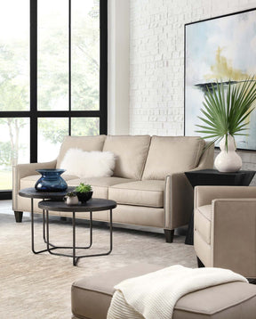 Lifestyle of a tan leather couch against a white brick wall and floor to ceiling windows adjacent to it.