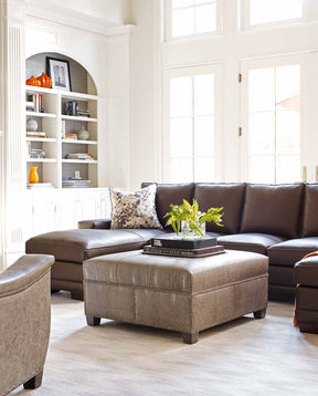 Lifestyle of a dark brown leather sectional with a lighter brown ottoman in front with a white built in bookshelf to the side of it