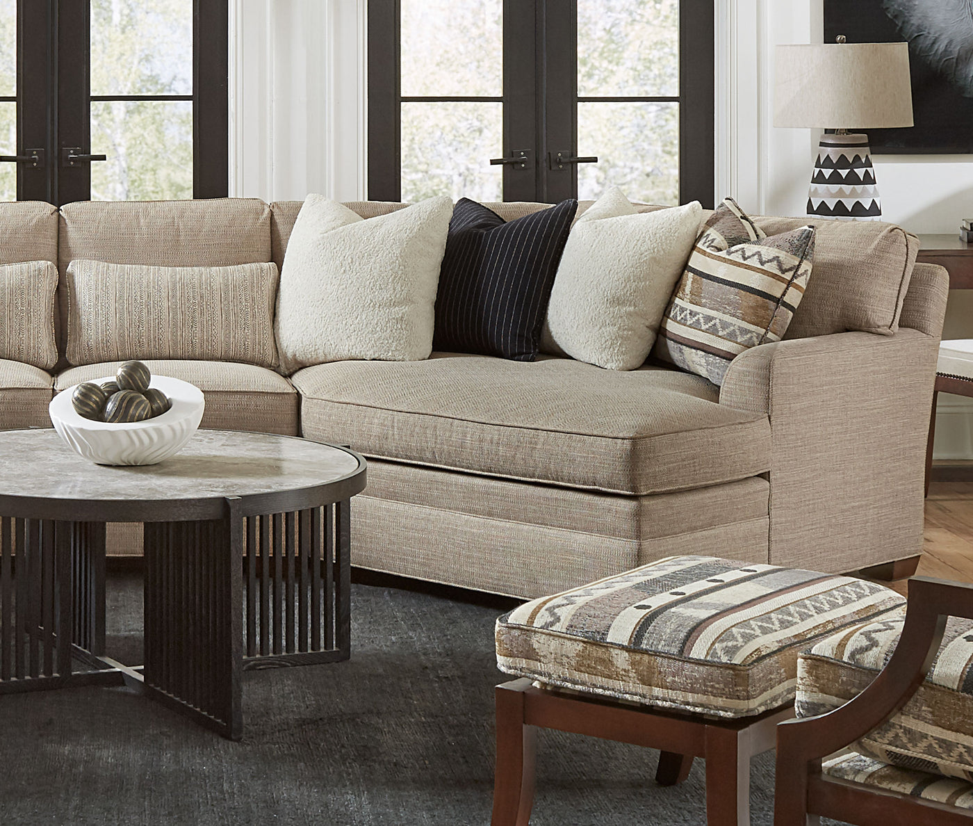 A 7000 Series sectional with light tan colored fabric, the sectional has many white, black, or tan and white decorative pillows, a Park Slope Round Cocktail Table sits in front of the table 