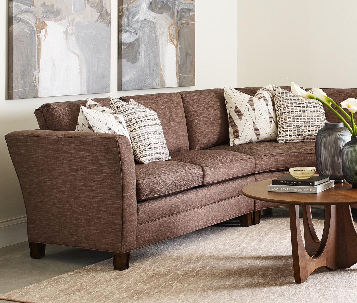 A dark brown fabric sectional in the corner of a cream-colored room with a Walnut Grove Round Cocktail Table in front of it.