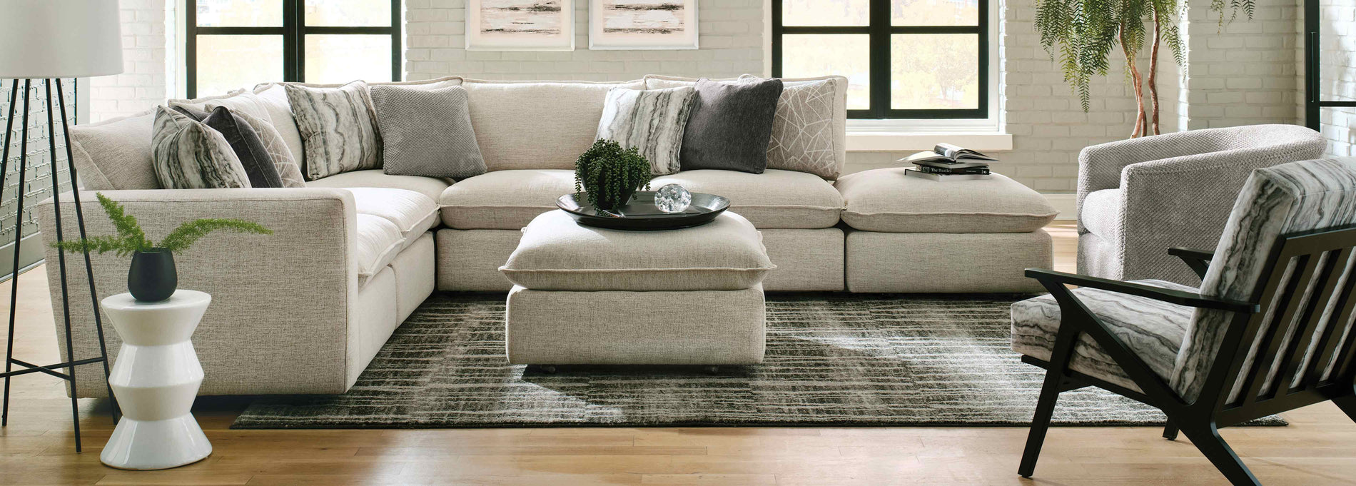 Lifestyle of a light gray sectional and matching ottoman sitting against a white wall and two windows