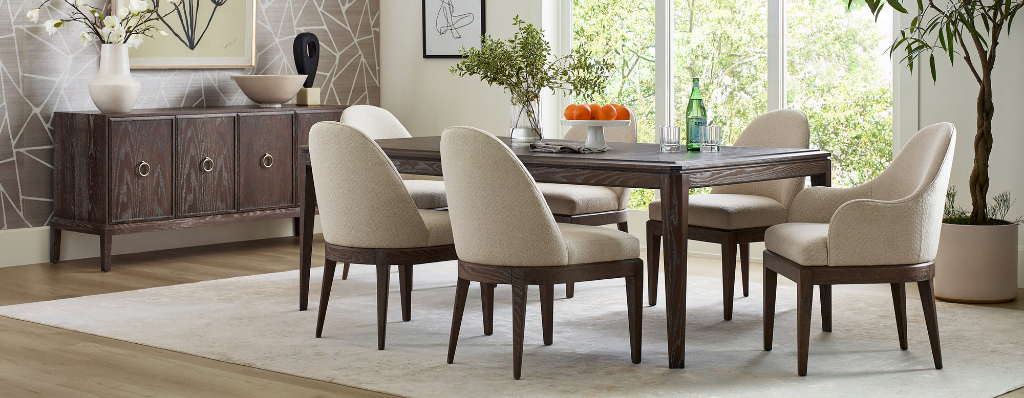 A Maidstone Rectangular Dining Table is in the middle of a brightly lit room surrounded by two Maidstone Upholstered Arm Chalirs and four Maidstone Upholstered Side Chairs. There is a Maidstone Four-Door Server in 202 Pier in the background.
