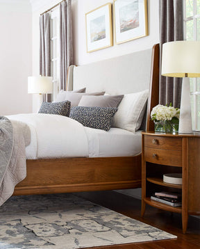 A Martine Bed with Upholstered Headboard sits against a white bed with a Martine Oval Nightstand beside it, there is a lamp and small white floral bouquet on the nightstand 