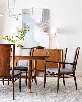 A Martine Dining Table is surrounded by Martine arm and side chairs, there is a Martine Sideboard in the background with a light gray and white painting resting above it against a white wall
