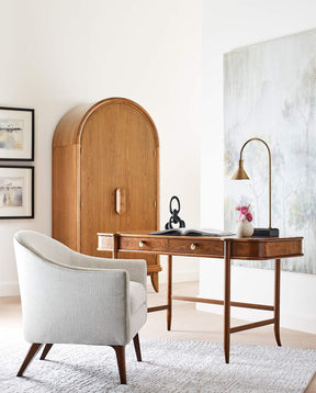 A Martine Vanity Desk is in a white room, with a gray upholstered chair slightly pulled out from it. There is a gold lamp and small vase on the desk and a Martine Arced Wardrobe in the background