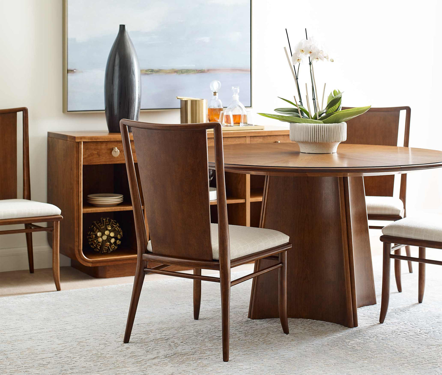 A Martine Sunburst Dining Table is surrounded by Martine Side Chairs, there is a Martine Server in the background with a drink tray and tall black vase on top of it
