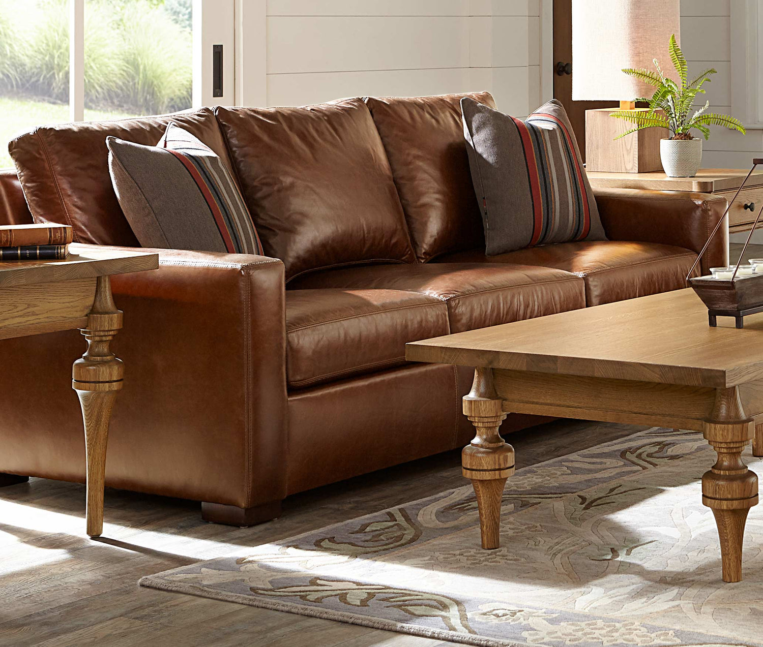 Lifestyle of a brown leather Memphis Sofa with two striped decorative pillows against both arms. There is a St. Lawrence Turned Cocktail Table in front of it, and a matching end table on either side