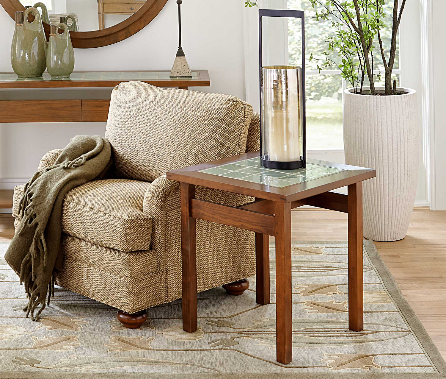 A Mission Tile-Top End Table sits next to a tan fabric accent chair that has a dark green throw blanket over the arm. There is a glass vase on top of the end table