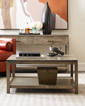 A Dwyer Square Coffee Table in the color 803-BLUFF sits in front of a red leather Paxton Chair and pink upholstered Paxton Sofa. There is a matching Dwyer Console Table in the background.