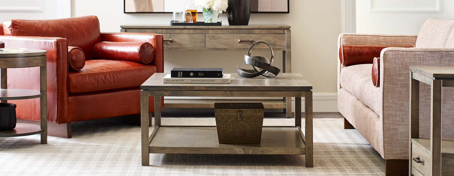 A Dwyer Square Coffee Table in the color 803-BLUFF sits in front of a red leather Paxton Chair and pink upholstered Paxton Sofa. There is a matching Dwyer Console Table in the background.