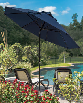 A large dark blue umbrella is open over two lounge chairs with a swimming pool in front of them and trees and a bright blue sky in the background