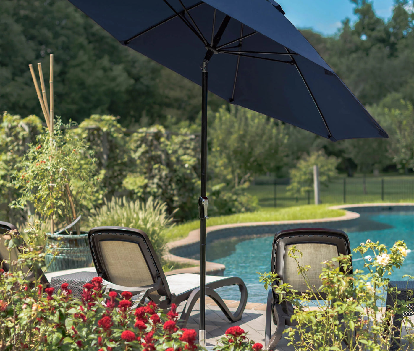 A Dark Blue Monterey 11' Crank Auto Tilt Outdoor umbrella is open above two lounge chairs that are sitting in front of a swimming pool. The pool and chairs are surrounded by a garden of plants and flowers