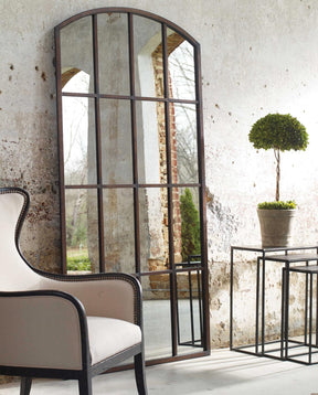An Uttermost Large Amiel Arch Mirror rests against a stone wall with a cream colored upholstered chair sat beside it