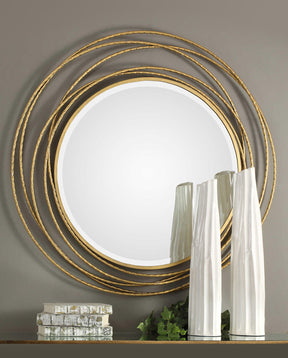 A round, gold framed mirror is mounted on a gray wall above a table that has decorative books and white vases on it