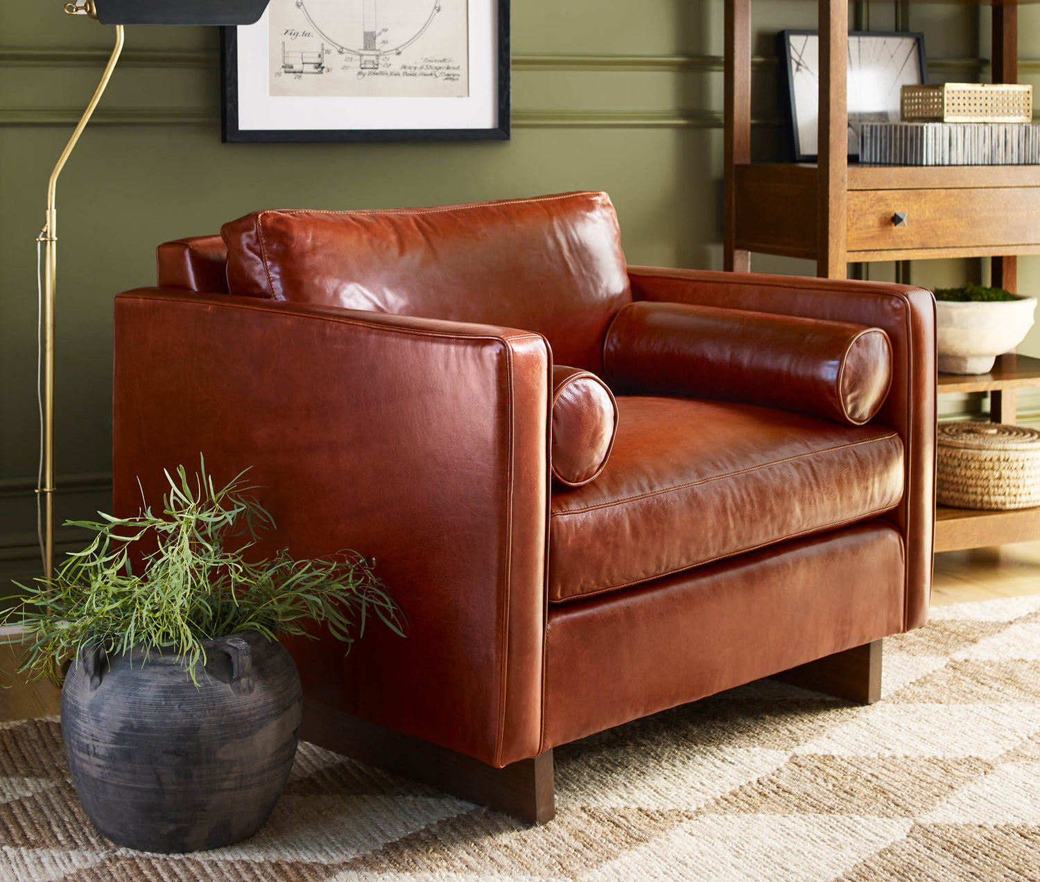 Brown Leather Paxton Sofa sitting in front of a dark green wall, there is a dark gray vase next to it on the floor with a plant inside of it and a gold floor lamp behind it