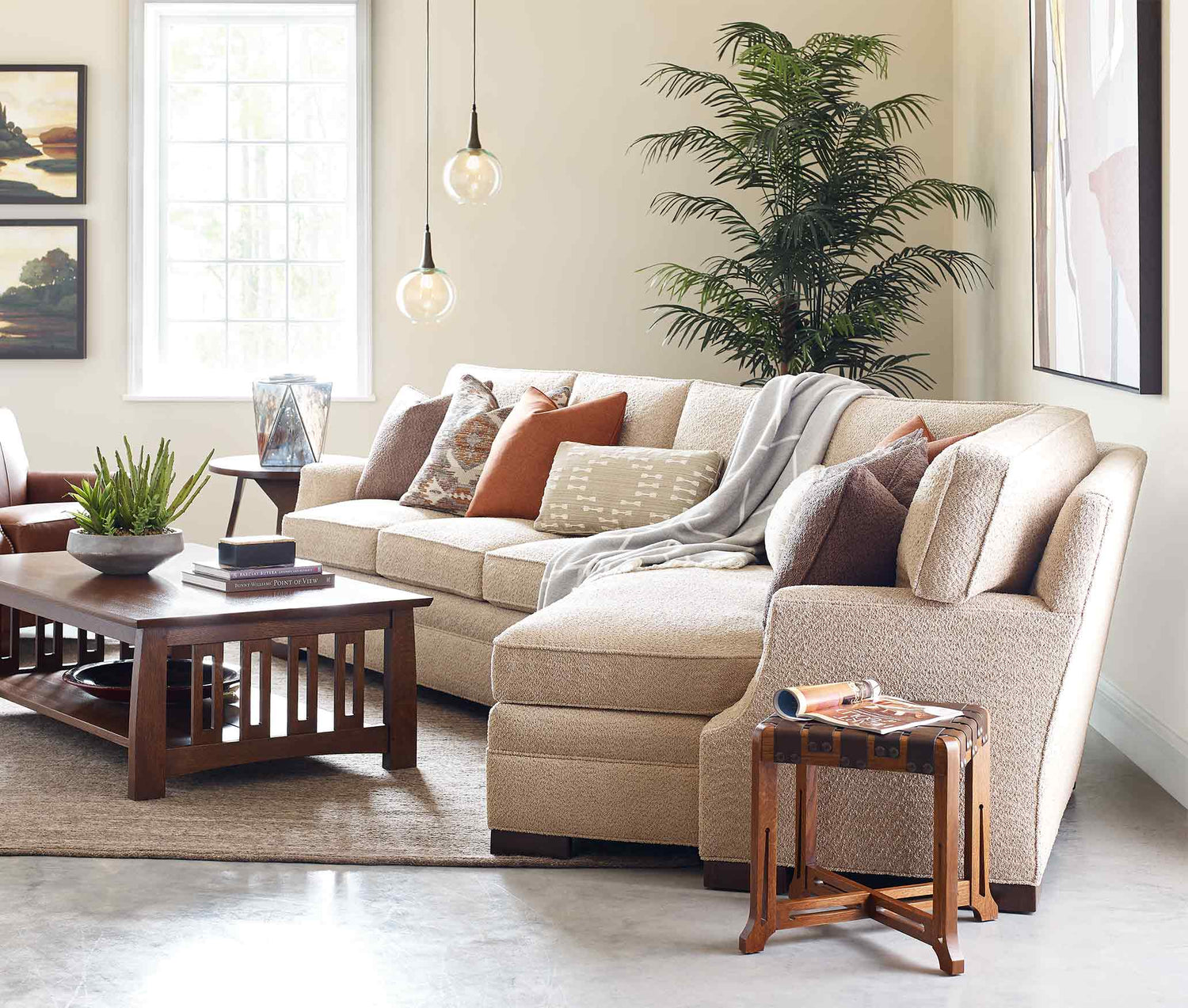 Lifestyle of a Cuddler shaped 7000 Series sectional that is light tan fabric. It has many accent pillows, and a throw blanket draped over the back of the sectional. There is a large fern behind the sectional against the white cream walls