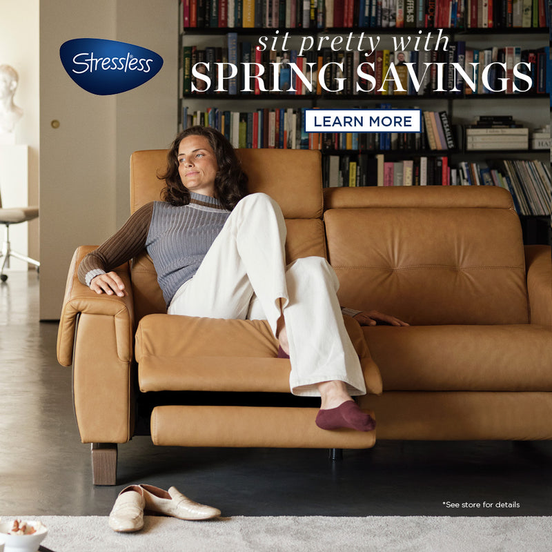 A square promotional image for Stressless featuring a woman lounging comfortably in a tan leather recliner. Behind her is a shelf full of books, suggesting a relaxing environment. The tagline 'sit pretty with SPRING SAVINGS' overlays the image at the top, while a 'LEARN MORE' button is positioned at the bottom. The promo hints at an elegant and comfortable lifestyle, encouraging viewers to take advantage of spring savings.