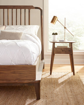 A lifestyle image of a Walnut Grove Spindle Bed that has a white comforter tucked into it and a Walnut Grove Nightstand next to it that has a gold lamp and small white vase on top of it