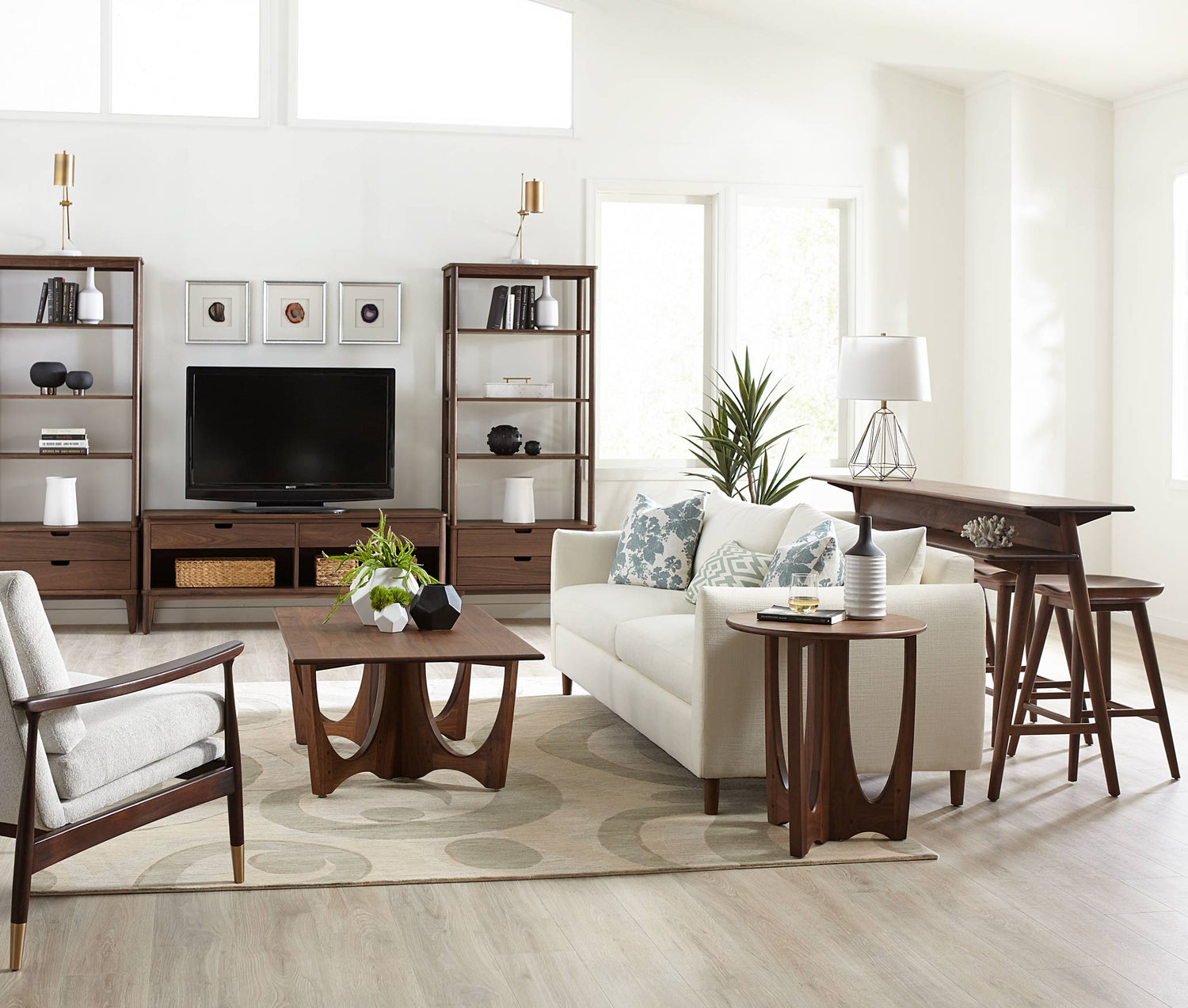 A Walnut Grove Morgan Sofa sits in a white, sunlight filled room with a matching gathering island with stools, end table, cocktail able surrounding it. There are two Walnut Grove Bookcases on either side of a Walnut Grove Entertainment Console that has a TV on it in the background