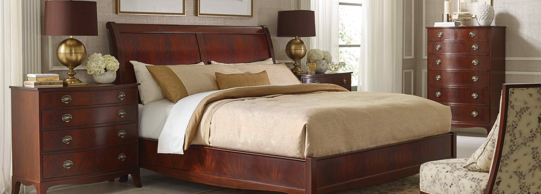 Lifestyle of a Whitehall Sleigh Bed that is against a light gray wallpapered wall, there are two Whitehall Four-Drawer Nightstand next to both sides of the bed with gold based lamps on top of them and a Whitehall Tall Chest in the background