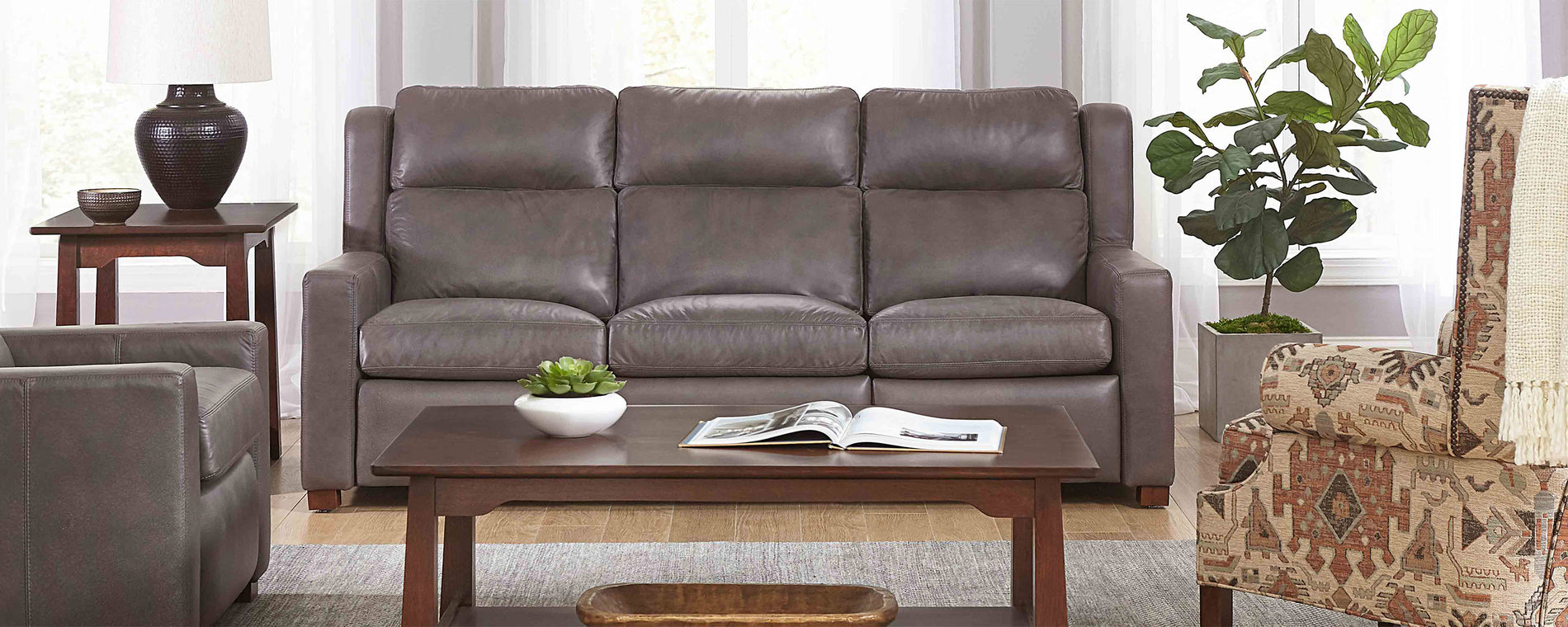 Lifestyle of a gray leather Woodlands Track Arm Sofa sitting against three tall windows, there is a matching Arm Recliner next to it and a dark wood coffee table in front of both