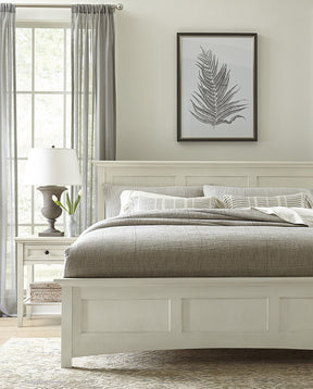 Origins by Stickley nighstand with a gray and white lamp on top in front of a floor to ceiling window with an Origins by Stickley white colored bed, there is a frame showing a drawn feather above the bed