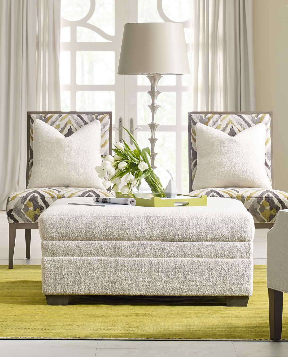 7000 Series white fabric ottoman that has a decorative tray with a small plant in it. The ottoman is sitting on top of a light green rug, with two fabric accent chairs behind it