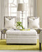 7000 Series white fabric ottoman that has a decorative tray with a small plant in it. The ottoman is sitting on top of a light green rug, with two fabric accent chairs behind it